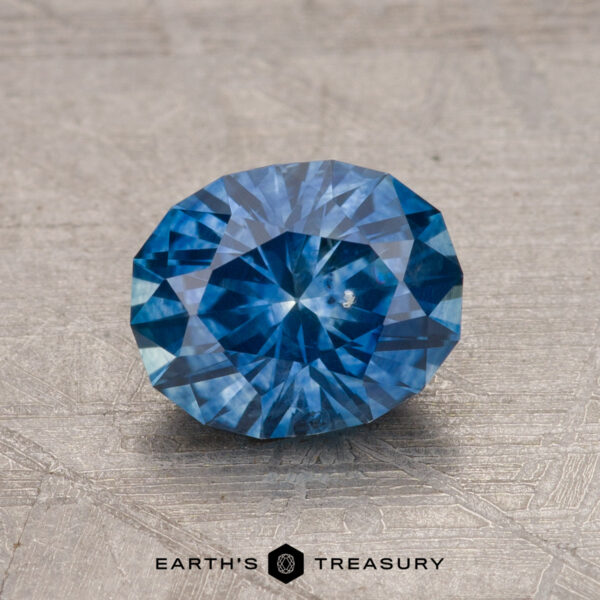 A faceted blue 1.45-carat Montana sapphire (heated) resting on a textured surface, with a logo reading "Earth's Treasury" at the bottom.