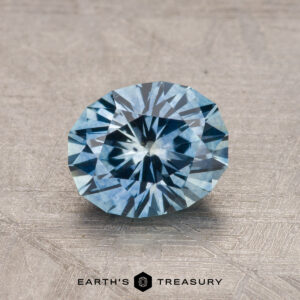 A 1.30-carat, round-cut Montana sapphire in a deep blue hue is showcased on a textured surface, revealing its intricate facets. The Earth's Treasury logo can be seen at the bottom.