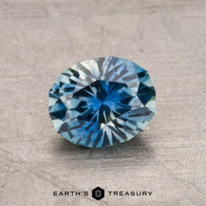 A faceted blue gemstone displayed on a textured surface, labeled "1.23-Carat Montana Sapphire (Heated)" at the bottom.