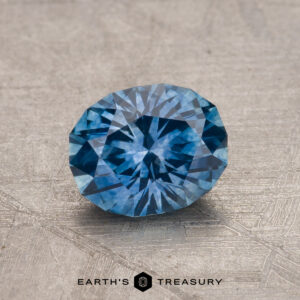 A round brilliant 1.21-carat Montana Sapphire gemstone, heated for enhanced quality, placed on a textured gray surface with the text "Earth's Treasury" beneath.