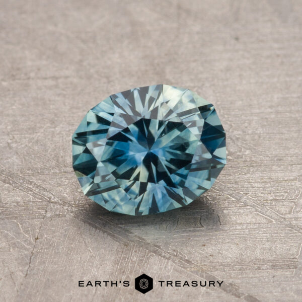 A faceted, round teal-blue 1.17-carat Montana sapphire is displayed on a textured gray surface, reflecting light to create intricate patterns. Text at the bottom reads "Earth's Treasury.