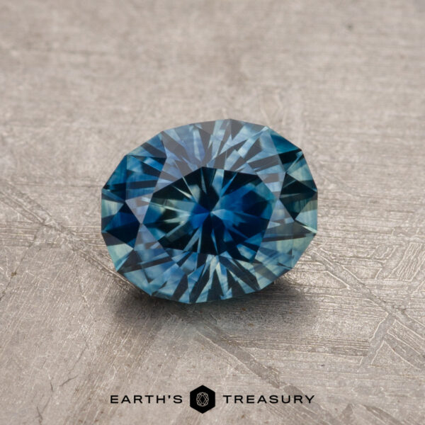 Faceted round blue gemstone with a textured surface, featuring the text "Earth's Treasury" at the base. Product Name: 1.15-Carat Montana Sapphire (Heated).