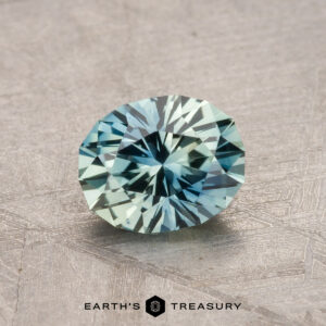 A 1.15-carat heated Montana sapphire with a blue-green hue rests on a textured surface.