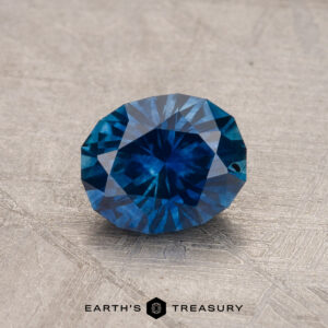 A faceted blue 1.14-carat Montana sapphire (heated) displayed on a textured surface with the logo "Earth's Treasury" at the bottom.