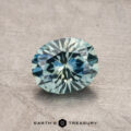 1.13-carat oval-cut Montana sapphire featuring a bluish-green hue, placed on a textured surface with the label "Earth's Treasury" at the bottom.