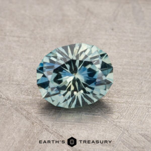 1.13-carat oval-cut Montana sapphire featuring a bluish-green hue, placed on a textured surface with the label "Earth's Treasury" at the bottom.