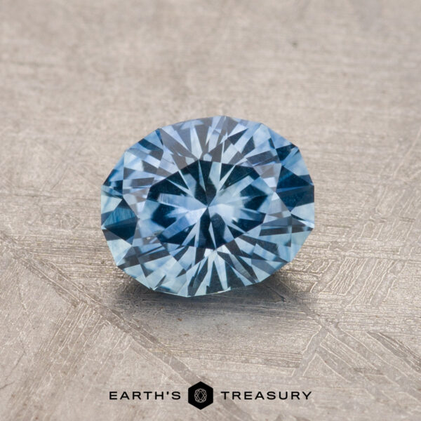 A 1.13-carat blue Montana Sapphire with intricate facets rests on a textured surface, featuring "Earth's Treasury" at the bottom.