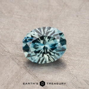 A 1.04-carat faceted blue-green Montana Sapphire rests on a textured surface, proudly displaying the "Earth's Treasury" brand at the bottom.