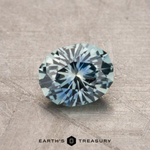 A faceted blue 1.00-carat Montana Sapphire (heated) is displayed on a textured surface with the text "Earth's Treasury" at the bottom.