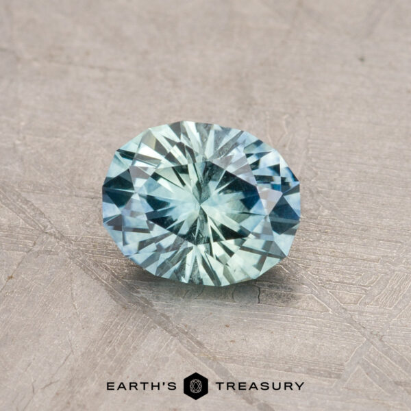 A faceted oval 0.95-carat Montana sapphire with a light blue hue rests on a textured gray surface. "Earth's Treasury" is written below.