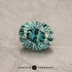 A round, faceted Montana sapphire weighing 0.94 carats sits on a textured surface, accompanied by the Earth's Treasury logo below.