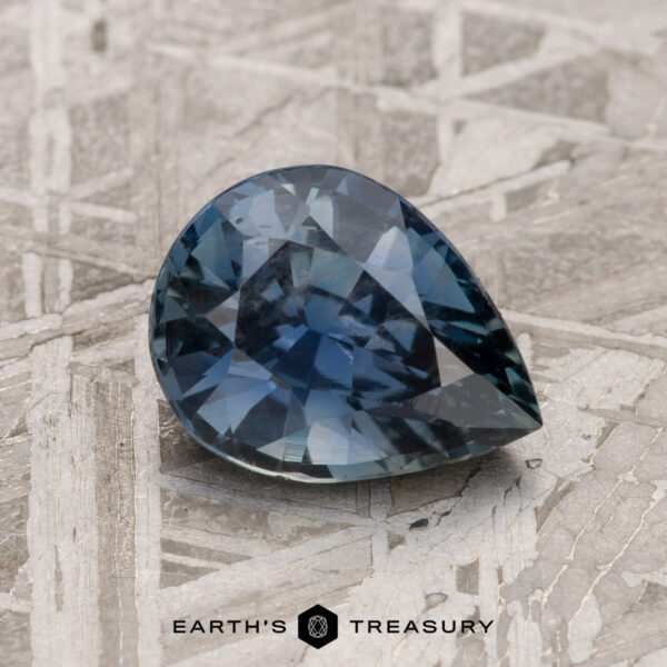 A 2.06-carat steely blue Tanzanian sapphire in a pear shape rests on a textured surface, accompanied by the "Earth's Treasury" logo below.