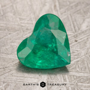 A heart-shaped green gemstone sits on a textured surface, showcasing the logo "Earth's Treasury" below. It is a 1.54-carat Colombian emerald that has been treated.