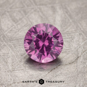 A 1.08-carat pastel pyrope garnet, round and faceted in a pink-purple hue, is showcased on a textured surface.