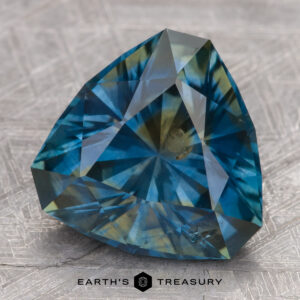 A 5.49-carat teal Montana sapphire with intricate trillion-cut facets on a textured surface, labeled "Earth's Treasury.