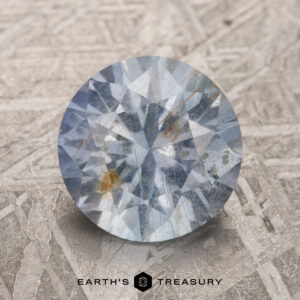 A round, faceted 2.65-carat Pale Gray-Blue Umba Sapphire featuring light blue hues with orange inclusions, set against a textured gray surface, and displaying the Earth's Treasury logo at the bottom.