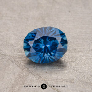 A 0.91-carat Montana Sapphire (heated), featuring a round faceted cut and blue hue, is showcased on a textured gray surface with the text "Earth's Treasury" below.