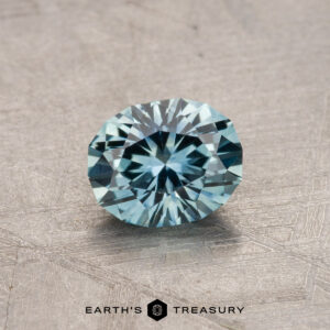 A round, faceted blue Montana Sapphire resting on a textured surface with the "Earth's Treasury" logo at the bottom.