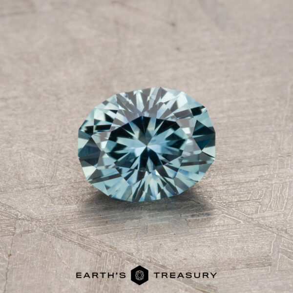 A round, faceted blue Montana Sapphire resting on a textured surface with the "Earth's Treasury" logo at the bottom.