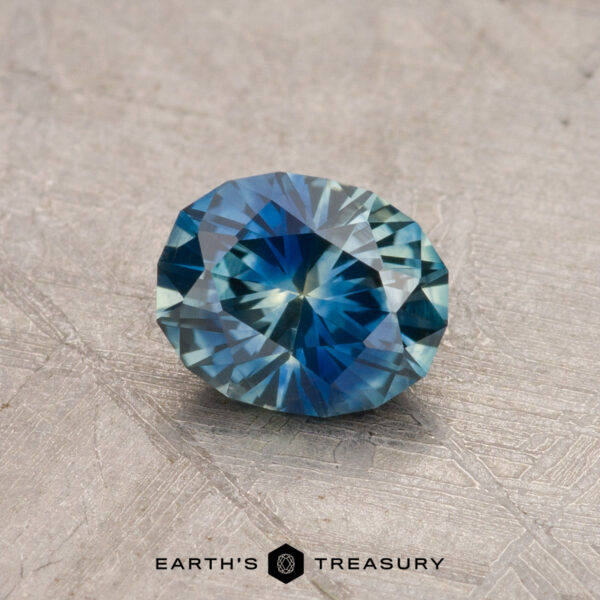 A faceted, blue-green oval 0.87-carat Montana sapphire (heated) resting on a textured gray surface, adorned with the "Earth's Treasury" logo at the bottom.