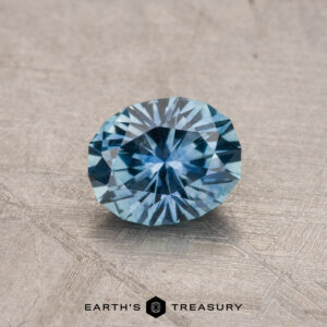 A round, faceted 0.81-carat Montana Sapphire with a blue hue is placed on a textured, light gray surface. The image features the text "Earth's Treasury" at the bottom.