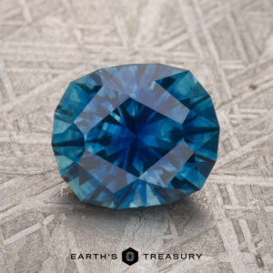 A 2.77-carat peacock blue Montana sapphire with intricate facets and a rectangular shape rests on a textured surface.