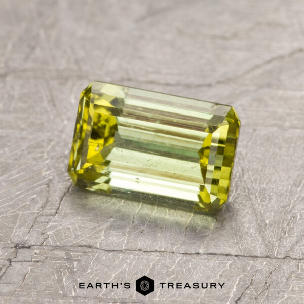 The ET1443 gemstone, featuring a faceted cut, sits on a textured gray surface in a rectangular green form.