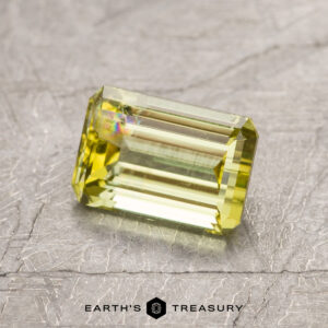 Rectangular, emerald-cut yellow gemstone with facets reflecting light, placed on a textured gray surface. Text "ET1447" visible on the bottom.