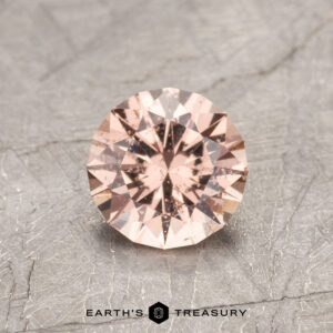 The ET1515, a round, faceted peach gemstone, is showcased on a textured gray surface.