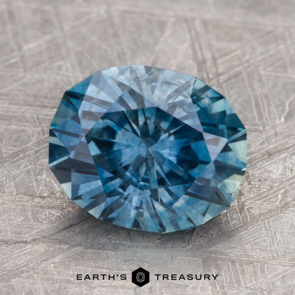 Close-up of a round, faceted teal blue gemstone on a textured gray surface. The "3.12-Carat Teal Blue Montana Sapphire (Heated)" by Earth's Treasury logo is visible at the bottom.