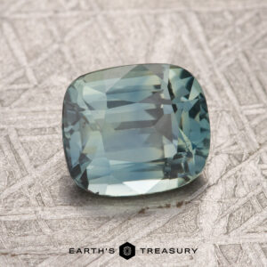 A 2.07-carat grayish blue-green Songea sapphire rests on a textured surface, with "Earth's Treasury" printed at the bottom.