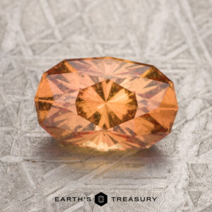 A 2.53-carat orange Umba sapphire is displayed on a textured metallic surface, with the "Earth's Treasury" logo below.