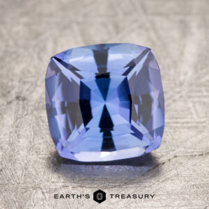 A 2.38-carat unheated tanzanite (zoisite) with a cushion-cut and faceted surfaces is displayed on a textured metal surface, with the text "Earth's Treasury" visible at the bottom.