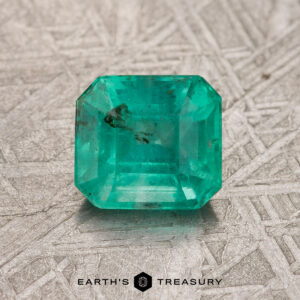 A treated 1.14-carat Colombian emerald with a rich green hue, cut in an emerald shape, rests on a textured surface, featuring the words "Earth's Treasury" below it.