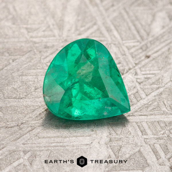 A heart-shaped Colombian emerald, weighing 0.86 carats and treated to enhance its beauty, rests on a textured gray surface with the inscription "Earth's Treasury" below.