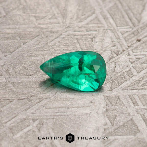 A 0.30-carat Colombian emerald (treated) in a pear shape is placed on a textured gray surface, with the "Earth's Treasury" logo at the bottom.