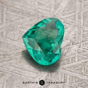 A heart-shaped emerald gemstone resting on a textured surface with the "Earth's Treasury" logo at the bottom, featuring a 0.77-carat treated Colombian emerald.