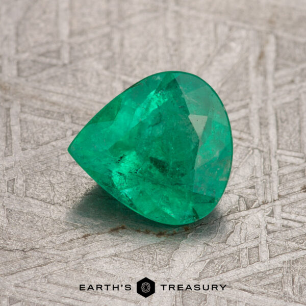 A 0.89-carat Colombian emerald with a triangular shape is set on a textured gray surface, and the words "Earth's Treasury" are visible below.