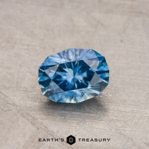 A 0.89-carat Montana Sapphire (heated), in blue, rests on a textured gray surface, with the "Earth's Treasury" logo displayed below.