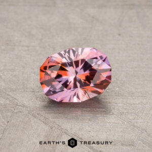 A 0.87-carat Montana sapphire, showcasing a faceted oval shape with pink and orange hues, is placed on a textured gray surface.