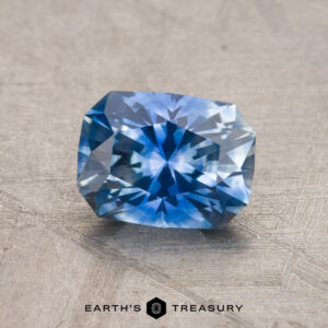 A 1.44-carat heated Montana Sapphire with a blue hue and a rectangular cushion cut, exhibiting intricate facets, is resting on a textured surface.