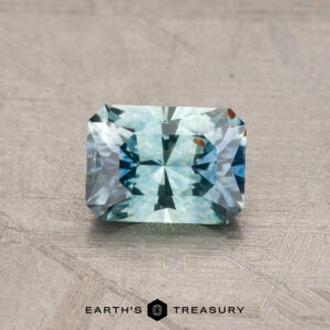 A rectangular, blue-green 1.06-carat Montana sapphire with facets rests on a textured gray surface. Logo text at the bottom reads: "Earth's Treasury.