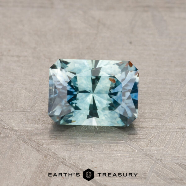 A rectangular, blue-green 1.06-carat Montana sapphire with facets rests on a textured gray surface. Logo text at the bottom reads: "Earth's Treasury.