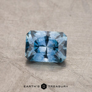 A rectangular faceted-cut blue gemstone, identified as a 0.81-carat Montana Sapphire (Heated), is showcased on a textured surface with the Earth's Treasury logo visible below.