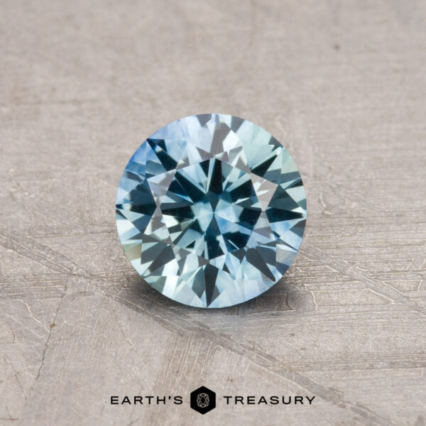 A 0.89-carat Montana sapphire, heated and round with blue-green facets, showcased on a textured surface.