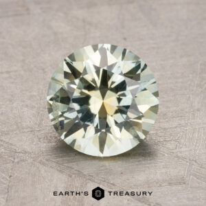 A round, faceted gemstone with a pale green hue is displayed on a textured surface. "1.79-Carat Montana Sapphire (Heated)" is printed below.