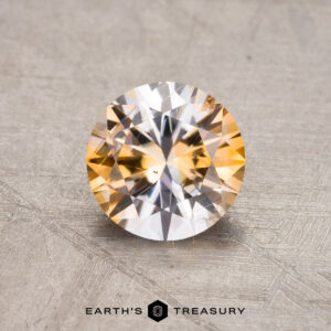 A round 1.21-carat Montana sapphire with a yellow-orange hue rests on a textured surface, with the words "Earth's Treasury" visible at the bottom.