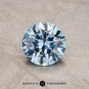 A 1.18-carat, round-cut Montana Sapphire (heated) in a light blue hue is displayed on a textured surface, with the logo "Earth's Treasury" beneath it.