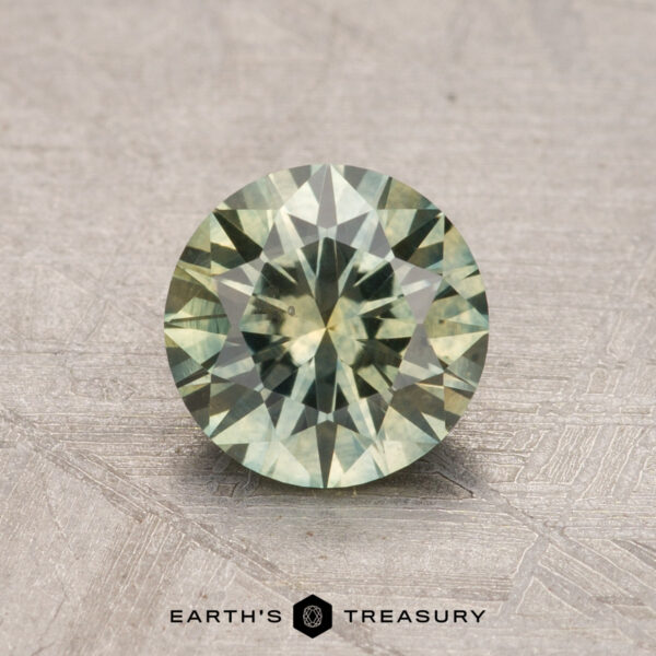 Round, faceted 1.18-carat Montana sapphire in a green shade set against a textured backdrop, displaying the "Earth's Treasury" logo beneath it.