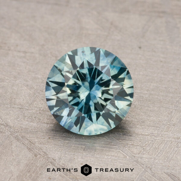 A round, faceted 1.14-carat Montana Sapphire with a blue hue is placed on a textured surface. The image includes the "Earth's Treasury" logo at the bottom.